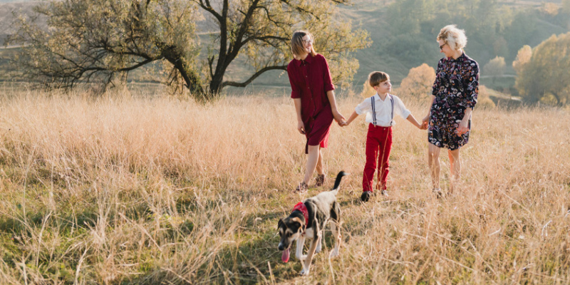 Two women holding hands with boy in grassy field, dog in foreground