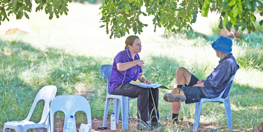 Woman talking to man whilst seated out doors with tree branch overhead.