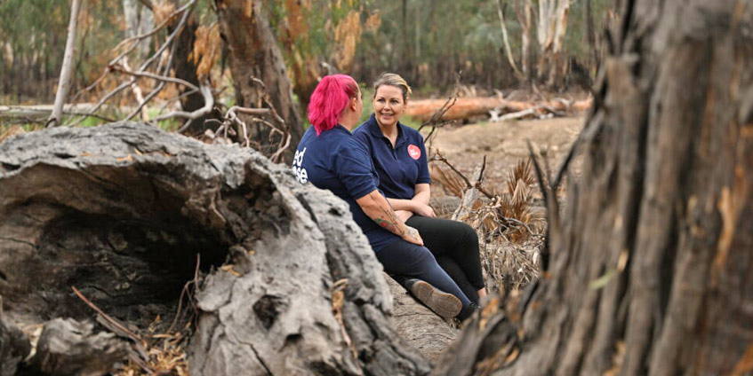 Skye with pink hair talking to a woman seated in the bush on some old fallen trees
