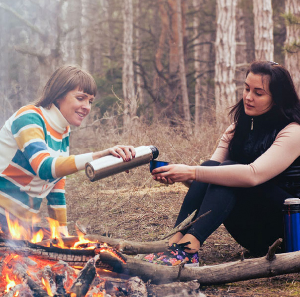 Two women having a warm drink near a campfire
