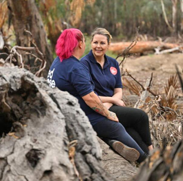Skye with pink hair talking to a woman seated in the bush on some old fallen trees